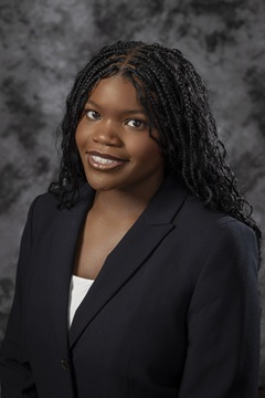 Professional studio portrait of a person with long braided hair wearing a dark blazer over a light-colored top. The person is facing the camera against a gray mottled studio backdrop. The image is framed from the chest up and lit with even studio lighting.