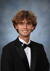 Formal studio portrait of a person with curly hair and eyeglasses wearing a black tuxedo jacket, white dress shirt, and black bow tie. The person is facing the camera against a blue-gray mottled studio background. The image is framed from the chest up and lit with soft, even portrait lighting.