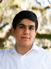 Head-and-shoulders portrait of a person wearing a white collared shirt, facing the camera. The image is taken outdoors with a softly blurred background of trees and greenery. The person has short, dark hair, and the lighting is natural and even. The composition centers the subject, with the upper torso visible and the background providing a warm, natural setting.
