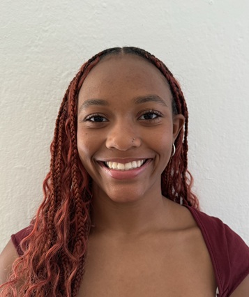 Head-and-shoulders portrait of a person with long, reddish braided hair wearing a burgundy top. The person is facing the camera and smiling, with a small nose stud and earrings visible. The background is a plain light-colored wall with soft, natural lighting.