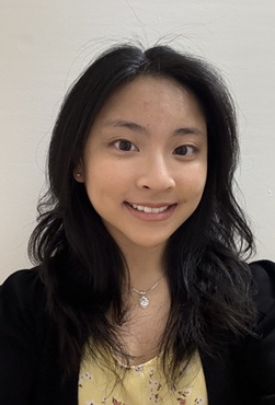Head-and-shoulders portrait of a person with shoulder-length dark hair wearing a black blazer over a light yellow patterned top. The person is facing the camera, wearing small stud earrings and a necklace with a pendant. The background is a plain light-colored wall with even indoor lighting.