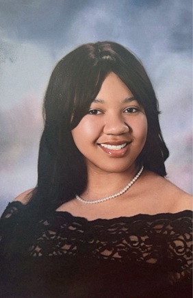 Formal studio portrait of a person with long, dark hair wearing a black off-the-shoulder lace top and a single-strand pearl necklace. The person is facing the camera against a soft blue-gray mottled studio background. The image is evenly lit and framed from the shoulders up.