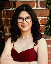 Professional portrait of a person with shoulder-length dark hair wearing rectangular eyeglasses and a sleeveless burgundy dress. The person is standing with arms crossed in front of a red brick wall. The image is framed from the waist up and lit with natural outdoor lighting.