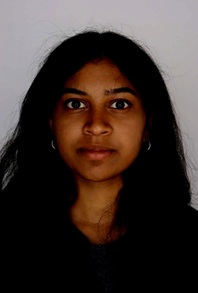 Head-and-shoulders portrait of a person with long, dark hair wearing a dark top and small hoop earrings. The person is facing the camera against a plain light-colored background. The image is closely cropped around the face and shoulders and lit with soft, even lighting, similar to an identification or school portrait photograph.