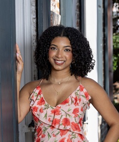 Outdoor portrait of a person with shoulder-length curly hair wearing a sleeveless floral dress with pink and coral flowers. The person is standing in a doorway with one hand resting on the doorframe and the other on their hip. Delicate necklaces are visible. The background includes a building entrance with glass panels and architectural details, softly blurred by the shallow depth of field. The image is framed from the waist up and lit with natural daylight.