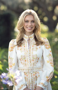 Outdoor portrait of a person with shoulder-length light-colored hair wearing a long-sleeved white dress with a floral pattern and decorative trim. The person is standing with hands clasped in front, facing the camera in a garden setting. Softly blurred greenery, flowers, and trees fill the background. The image is framed from about the knees up and lit with natural daylight, creating a bright, professional portrait.