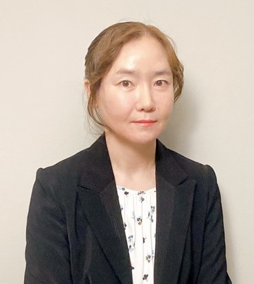 A person with brown hair pulled back is shown from the chest up against a plain tan background. They are wearing a black blazer and a white top with small flowers underneath. Their posture is relaxed.