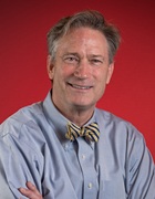 Professional studio portrait of a person wearing a light blue button-down shirt and a patterned bow tie. The image is framed from the chest up against a solid red background. The person is posed with arms crossed and facing the camera. The portrait is evenly lit and presented in a professional academic or business style.