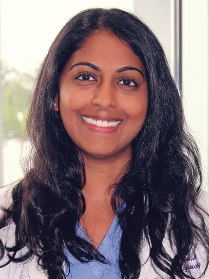 Professional head-and-shoulders portrait of a person wearing a white medical coat over light blue clinical scrubs. The person has long dark hair and is facing the camera. The image is photographed indoors with large windows in the background, which are softly out of focus. The portrait is well lit and framed from the chest up in a clinical or academic professional setting.