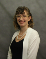 Professional head-and-shoulders portrait of a person with curly shoulder-length hair and glasses, wearing a black top, white cardigan, and layered pearl necklace. The person is seated slightly angled toward the camera against a gray studio backdrop.