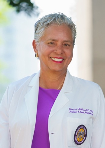 Professional portrait of a person standing outdoors against a softly blurred background of light-colored architectural columns and greenery. The person is wearing a white medical coat over a bright purple top, with embroidered text and a university-style emblem visible on the coat pocket. The image is well lit with natural light and framed from about the waist up, with the person facing the camera in a clinical or academic setting.
