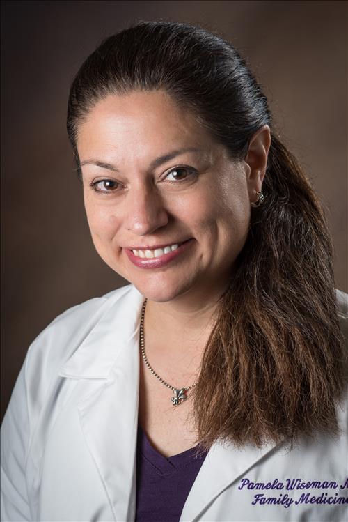 Professional head-and-shoulders portrait of a person wearing a white medical coat over a dark purple top. The coat features embroidered text on the lower right side, partially visible and including the words “Family Medicine.” The person is also wearing a necklace and small earrings. The image is set against a softly lit brown studio background and is framed from the shoulders up in a clinical or academic professional style.