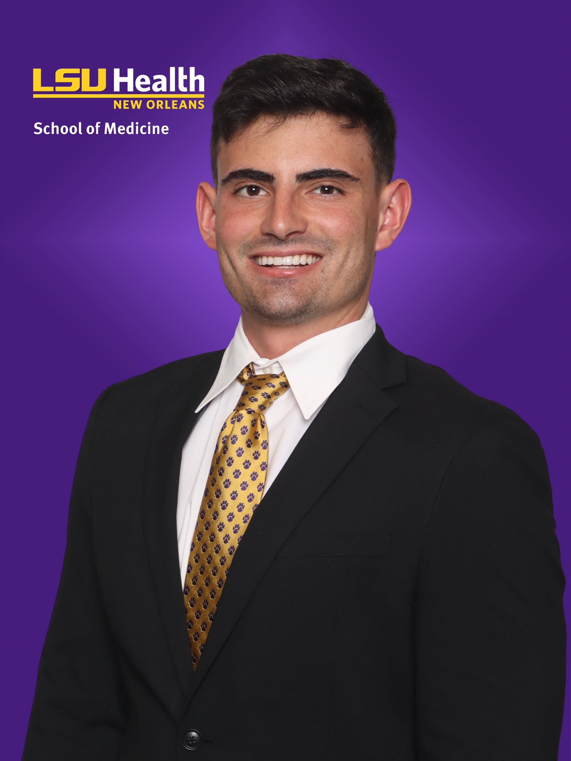 Professional school-affiliated headshot showing a person in business attire, including a dark suit and gold patterned tie, posed against a purple LSU Health New Orleans School of Medicine branded background. Visible text in the upper left reads “LSU Health New Orleans” and “School of Medicine.”