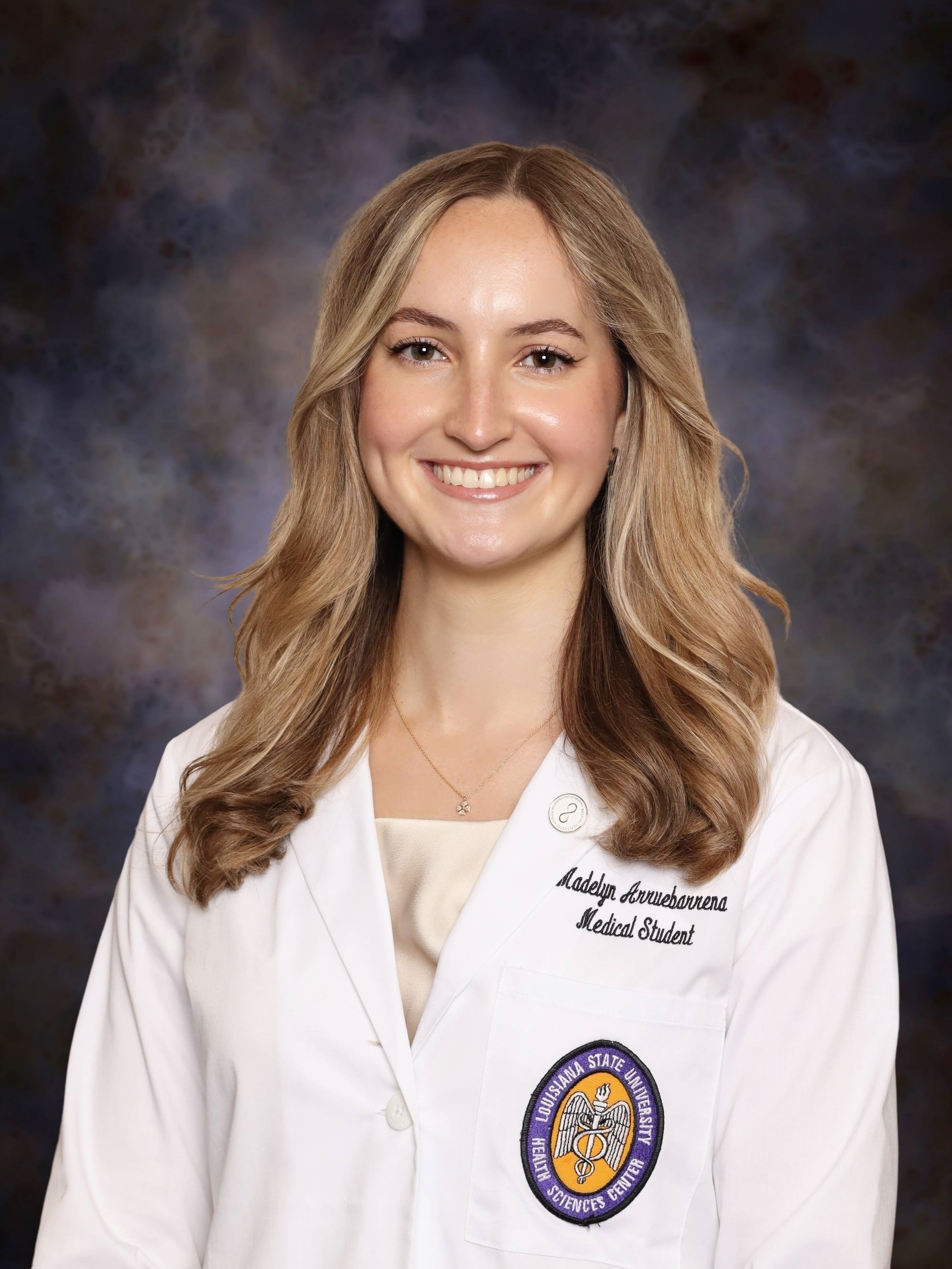 Professional medical school headshot showing a person in a white medical coat with a Louisiana State University Health Sciences Center emblem on the chest pocket. Visible embroidered text on the coat includes the person's name and the title “Medical Student.” The image is evenly lit with a neutral studio background that keeps attention on the subject.