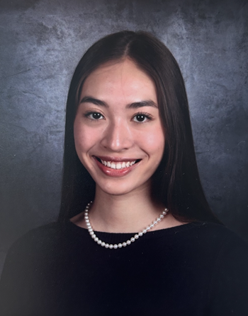 Formal head-and-shoulders portrait of a person with long, straight dark hair, wearing a dark top and a single strand of pearls. The person is facing the camera and centered against a mottled gray studio background. The image is evenly lit and cropped from the upper chest upward, resembling a professional school or graduation-style portrait.