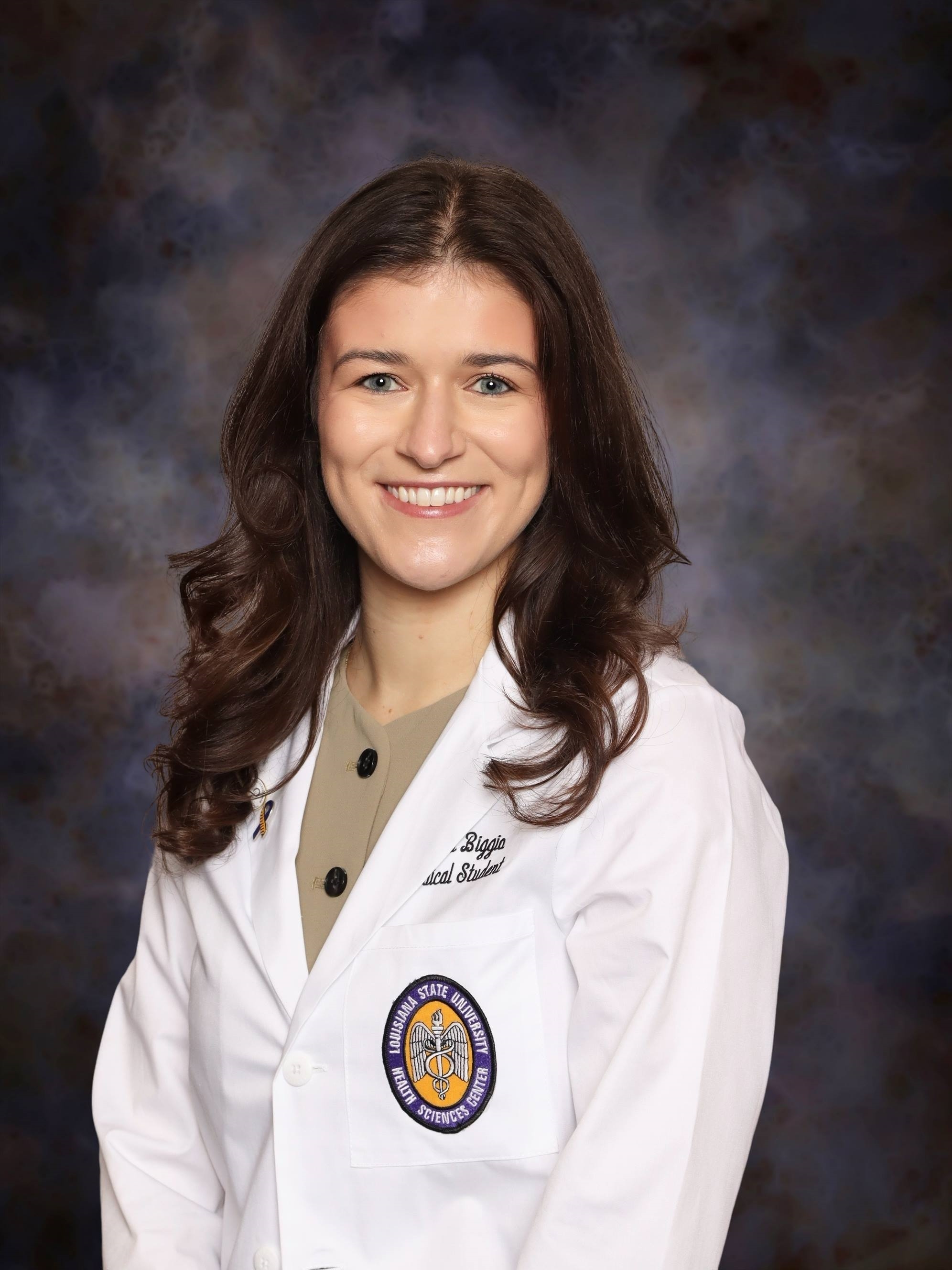 Professional medical school headshot showing a person wearing a white medical coat with embroidered text identifying the wearer as a medical student and a Louisiana State University Health Sciences Center emblem on the chest pocket. A small ribbon-shaped lapel pin is attached to the coat. The subject is wearing a light-colored top with dark buttons beneath the coat. The portrait is set against a dark blue and gray mottled studio background with even, professional lighting.