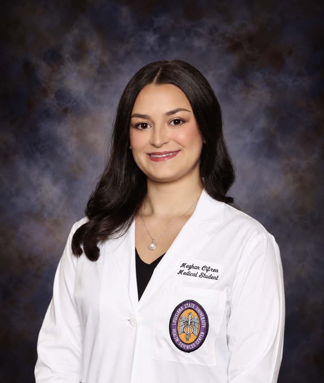 Professional studio headshot of a person standing against a dark blue and gray mottled backdrop. The person is wearing a white medical student coat over a dark top and a necklace with a round pendant. An embroidered Louisiana State University Health Sciences Center patch is visible on the left chest pocket of the coat. The portrait is framed from the chest up, with the person facing the camera directly. Embroidered text on the coat identifies the wearer as a medical student, and the lighting is even and professional, keeping the focus on the subject.