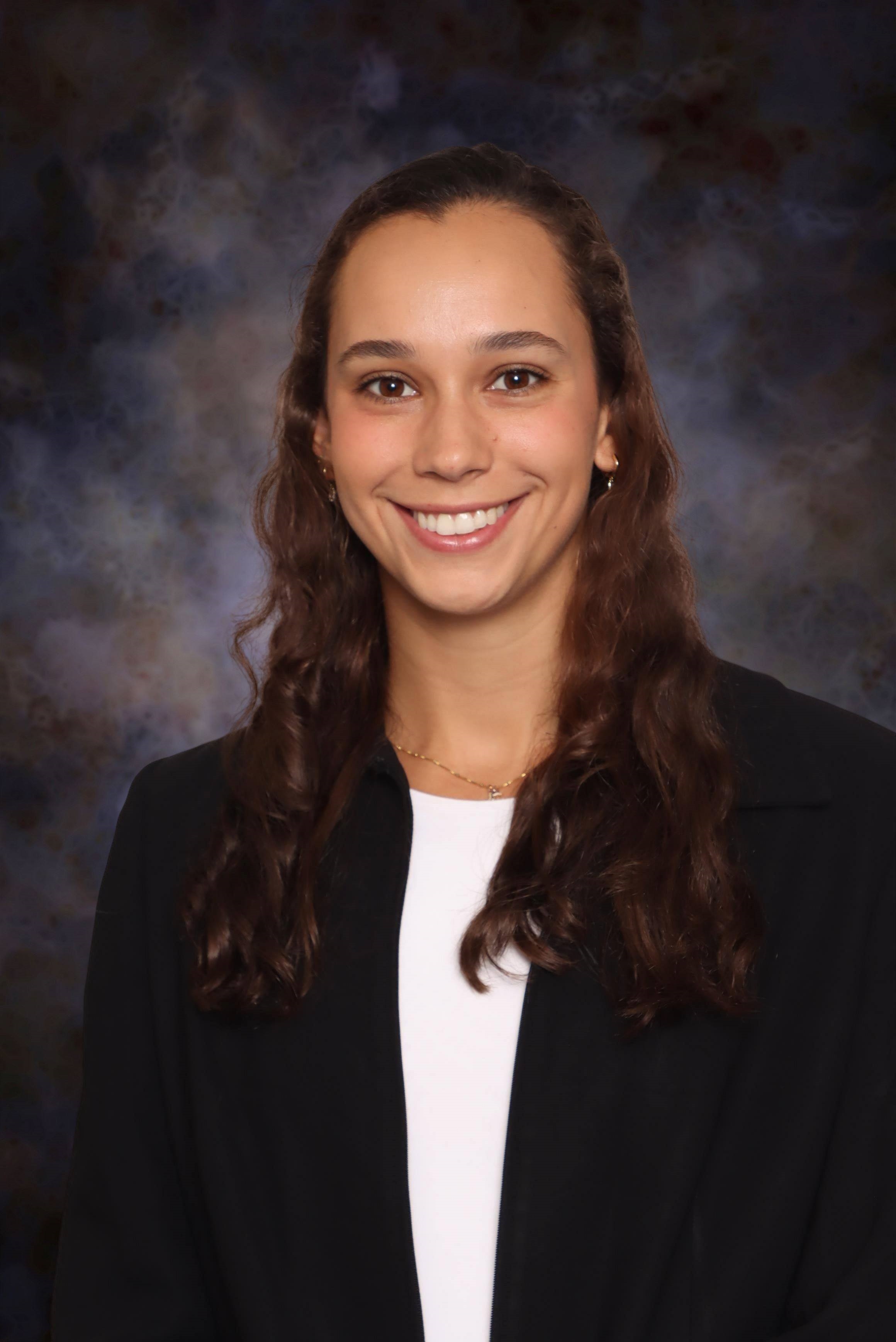 Professional headshot showing a person wearing a dark blazer over a light-colored top. The subject is facing the camera and wearing a delicate necklace and small earrings. The portrait is set against a dark blue and gray mottled studio background with even, professional lighting.