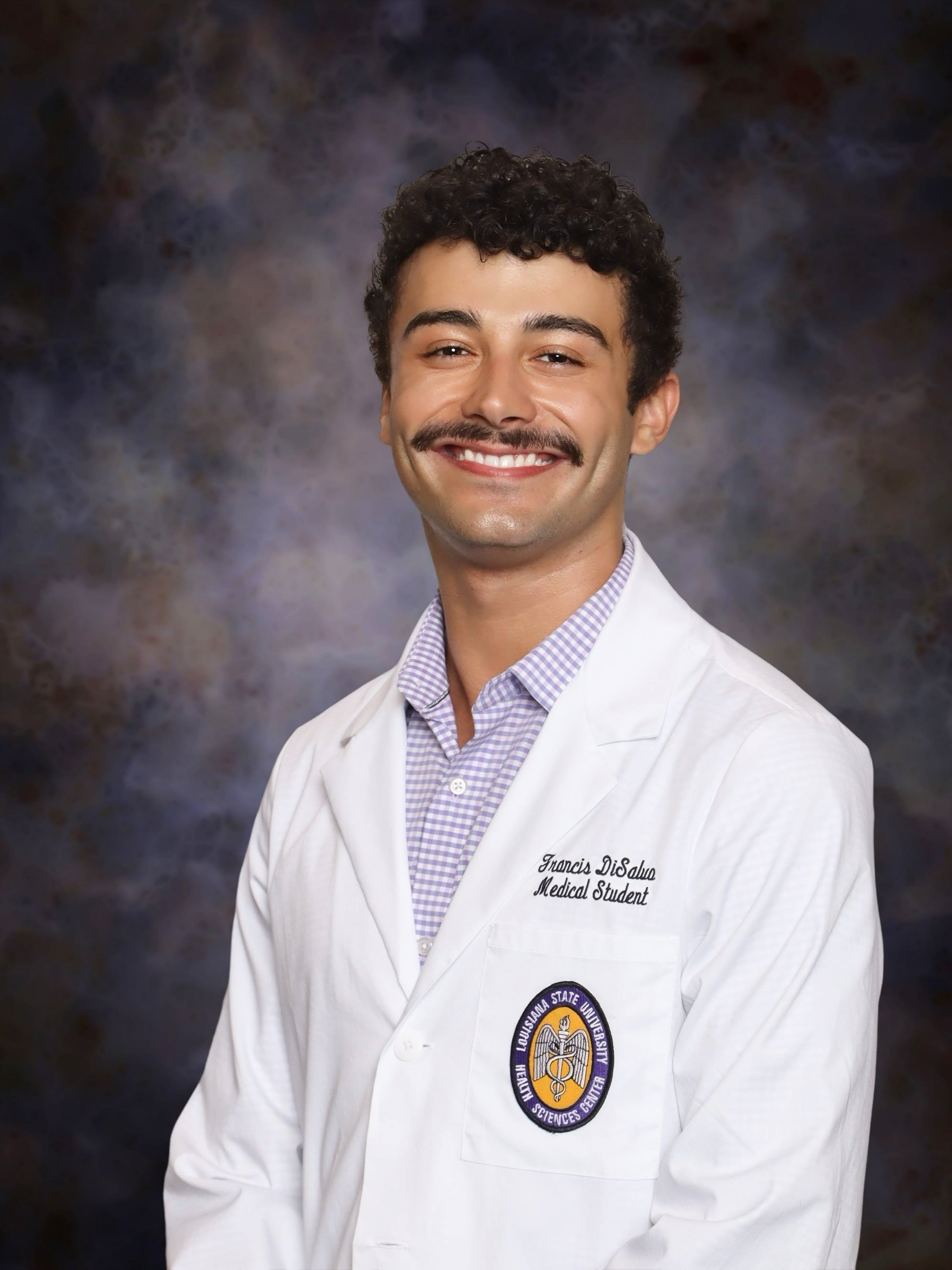 Professional medical school headshot showing a person wearing a white medical coat over a light purple checkered button-down shirt. Visible embroidered text on the coat includes the title “Medical Student,” and a Louisiana State University Health Sciences Center emblem is displayed on the chest pocket. The subject has curly hair and a mustache. The portrait is set against a dark blue and gray mottled studio background with even, professional lighting.