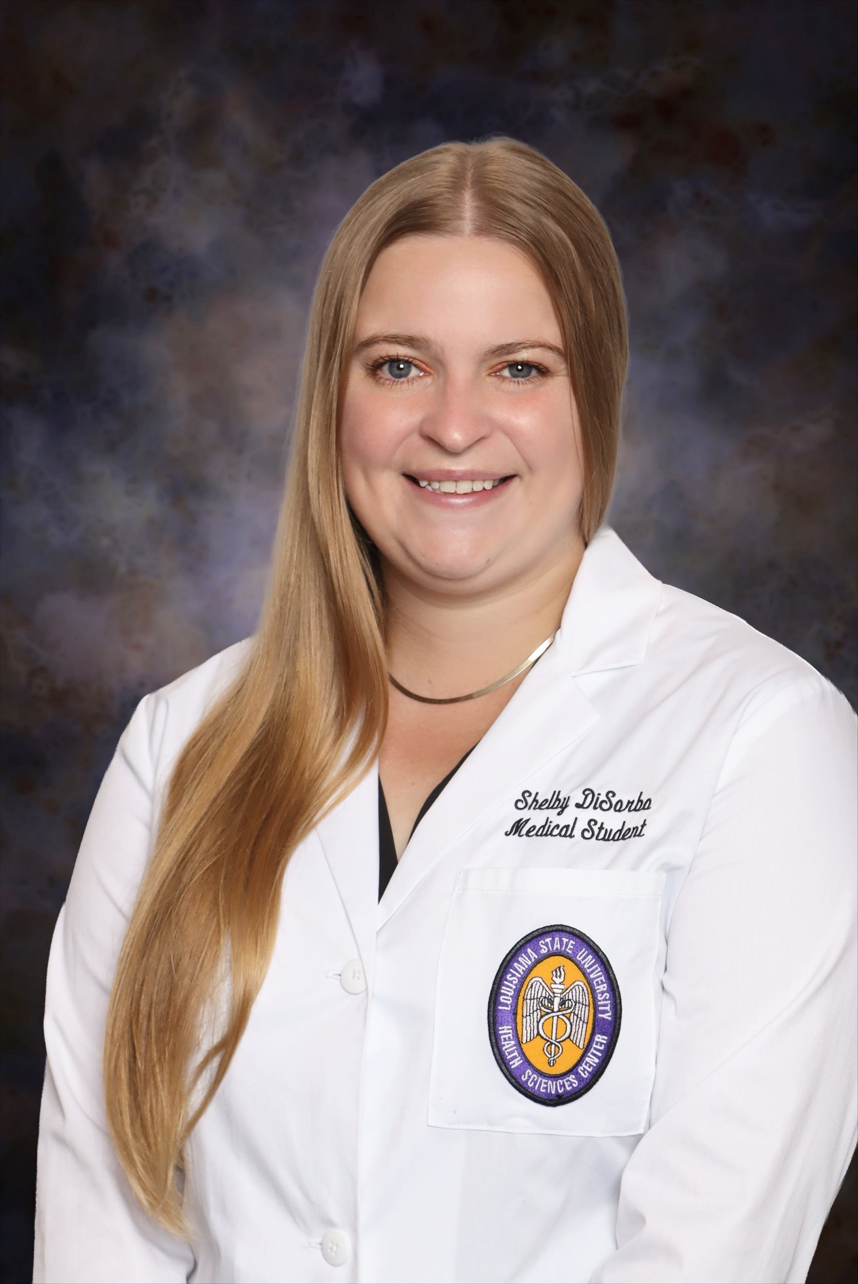 Professional medical school headshot showing a person wearing a white medical coat with embroidered text identifying the wearer as a medical student and a Louisiana State University Health Sciences Center emblem on the chest pocket. The subject is posed in front of a dark blue and gray mottled studio background and is wearing a dark top and a simple necklace. The image is evenly lit with the focus centered on the subject.