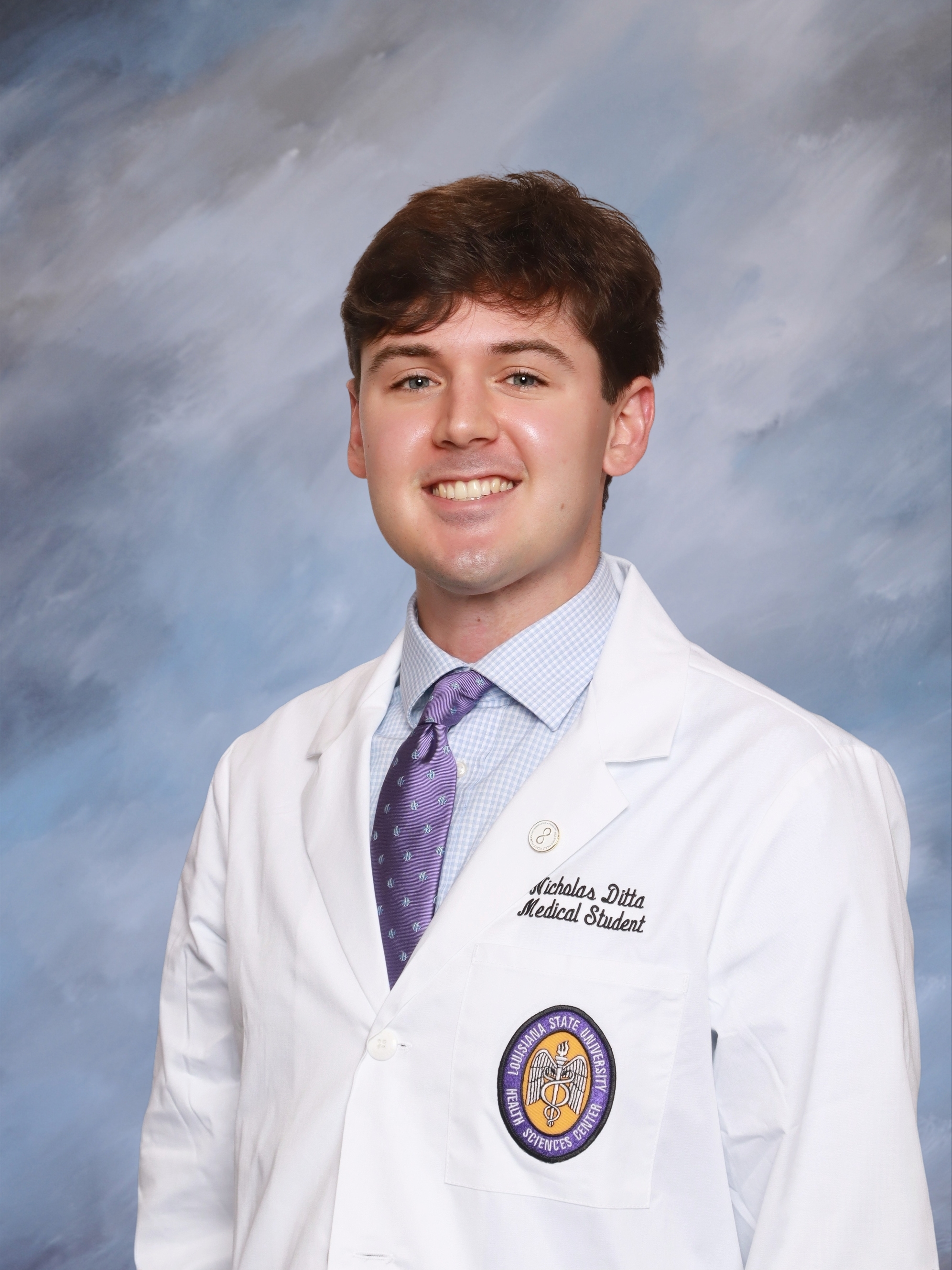 Professional medical school headshot showing a person wearing a white medical coat over a light blue dress shirt and a purple patterned tie. Visible embroidered text on the coat includes the title “Medical Student,” and a Louisiana State University Health Sciences Center emblem is displayed on the chest pocket. The subject is facing the camera against a softly textured blue studio background with even lighting.