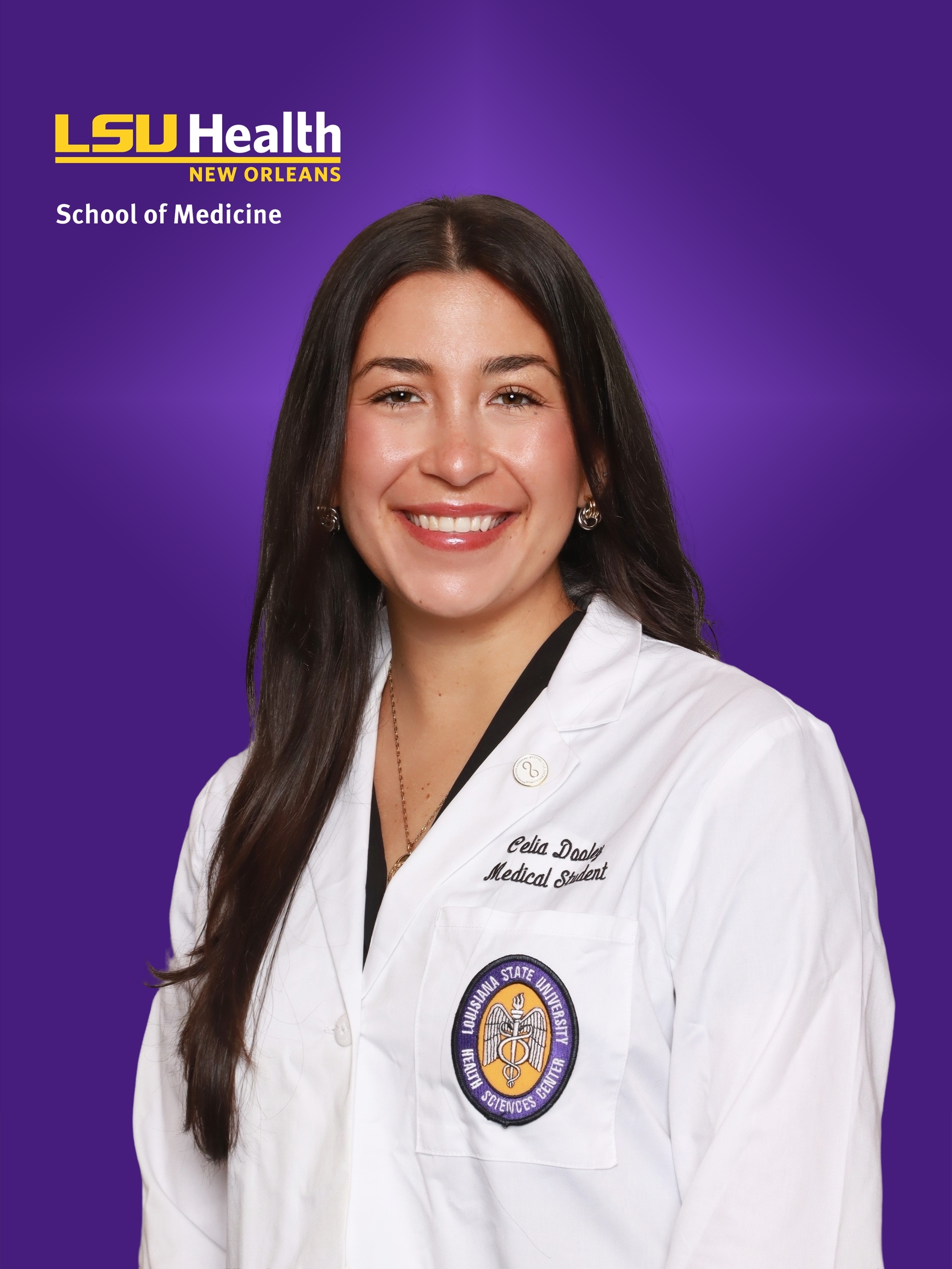 Professional medical school headshot showing a person wearing a white medical coat with embroidered text identifying the wearer as a medical student and a Louisiana State University Health Sciences Center emblem on the chest pocket. A small circular lapel pin is attached to the coat. The subject is wearing a dark top beneath the coat and small hoop earrings. The portrait is set against a purple LSU Health New Orleans School of Medicine branded background with the LSU Health New Orleans logo and “School of Medicine” text visible in the upper left corner.