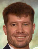 Professional headshot showing a person wearing a dark suit jacket, light-colored dress shirt, and patterned tie. The image is tightly cropped around the head and shoulders. The subject is facing the camera against a softly blended green and gray studio background with even, professional lighting.