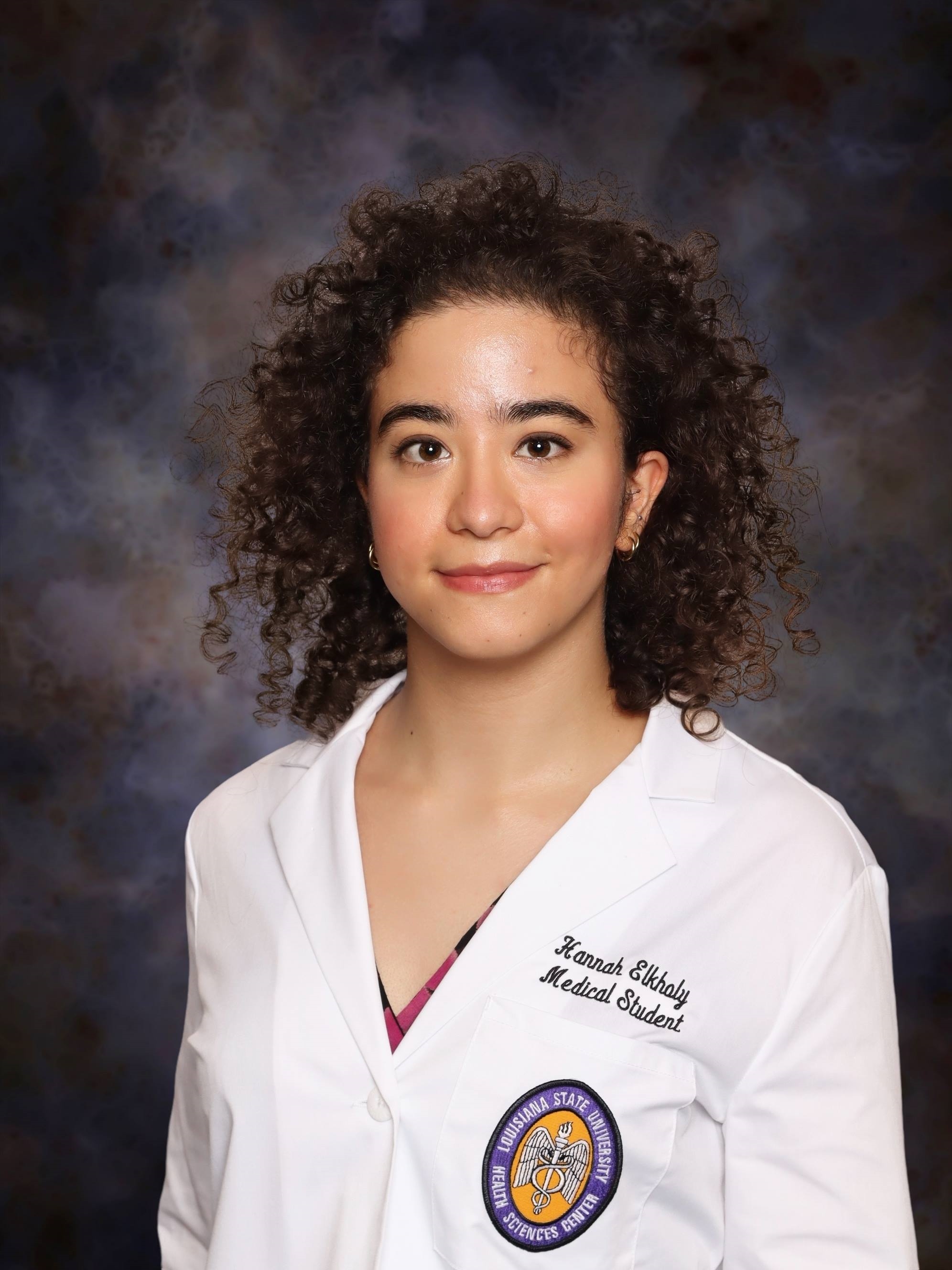 Professional medical school headshot showing a person wearing a white medical coat with embroidered text identifying the wearer as a medical student and a Louisiana State University Health Sciences Center emblem on the chest pocket. The subject is facing the camera and wearing a patterned top beneath the coat. The portrait is set against a dark blue and gray mottled studio background with even, professional lighting.