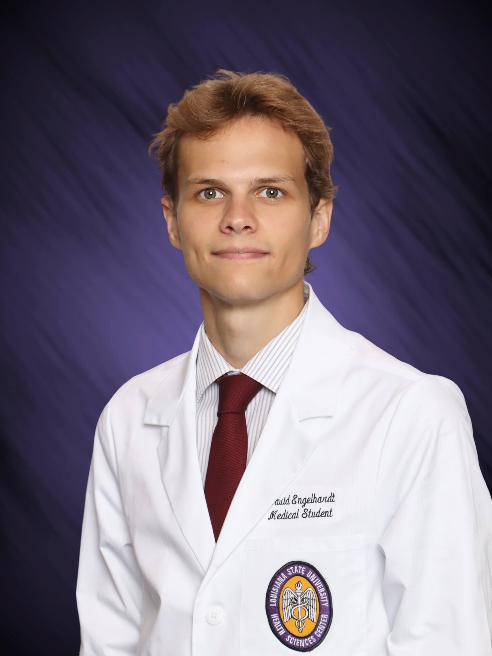 Professional medical school headshot showing a person wearing a white medical coat over a light-colored striped dress shirt and a dark red tie. Visible embroidered text on the coat includes the title “Medical Student,” and a Louisiana State University Health Sciences Center emblem is displayed on the chest pocket. The subject is facing the camera against a dark blue studio background with subtle diagonal texture and even, professional lighting.