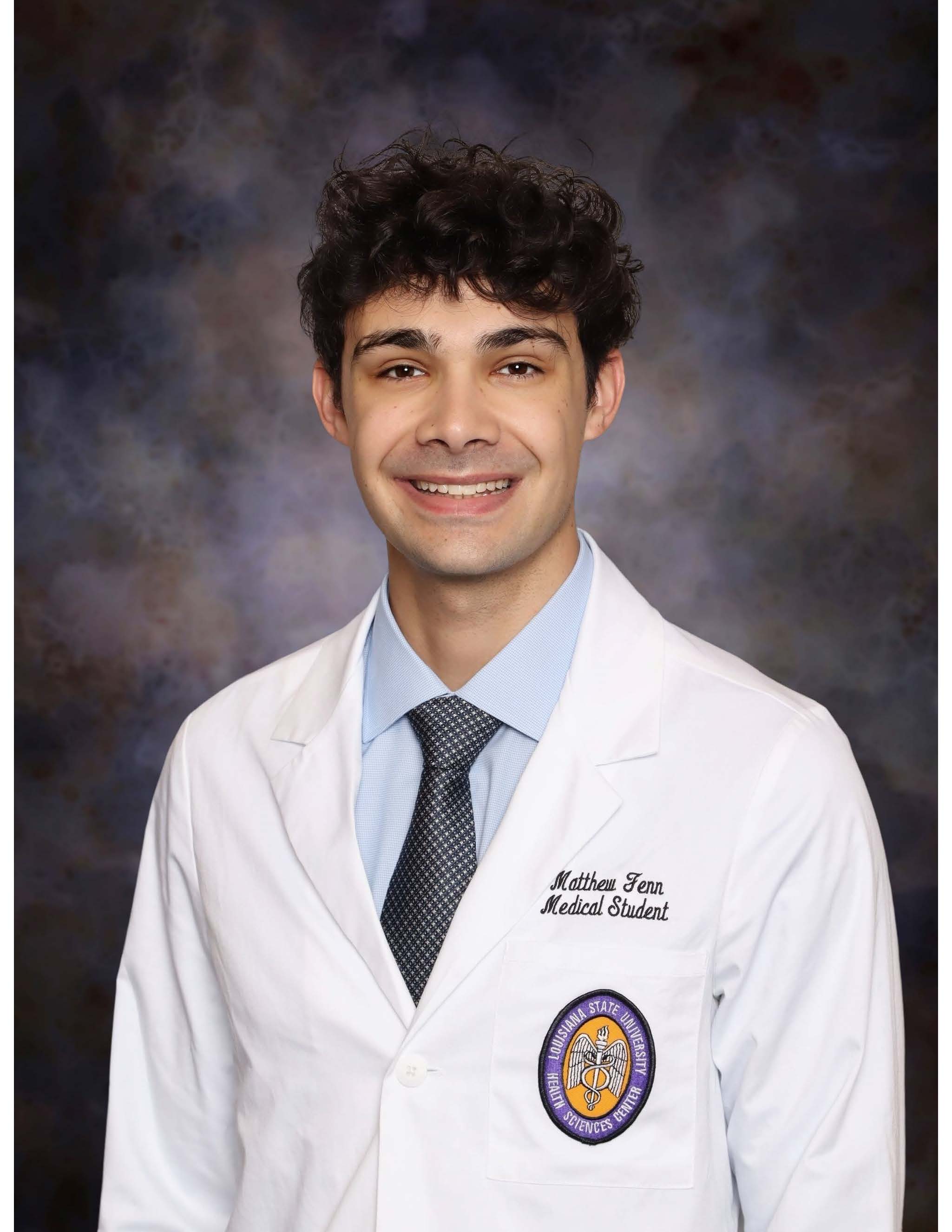 Professional medical school headshot showing a person wearing a white medical coat over a light blue dress shirt and patterned tie. Visible embroidered text on the coat includes the title “Medical Student,” and a Louisiana State University Health Sciences Center emblem is displayed on the chest pocket. The subject is facing the camera against a dark blue and gray mottled studio background with even, professional lighting.