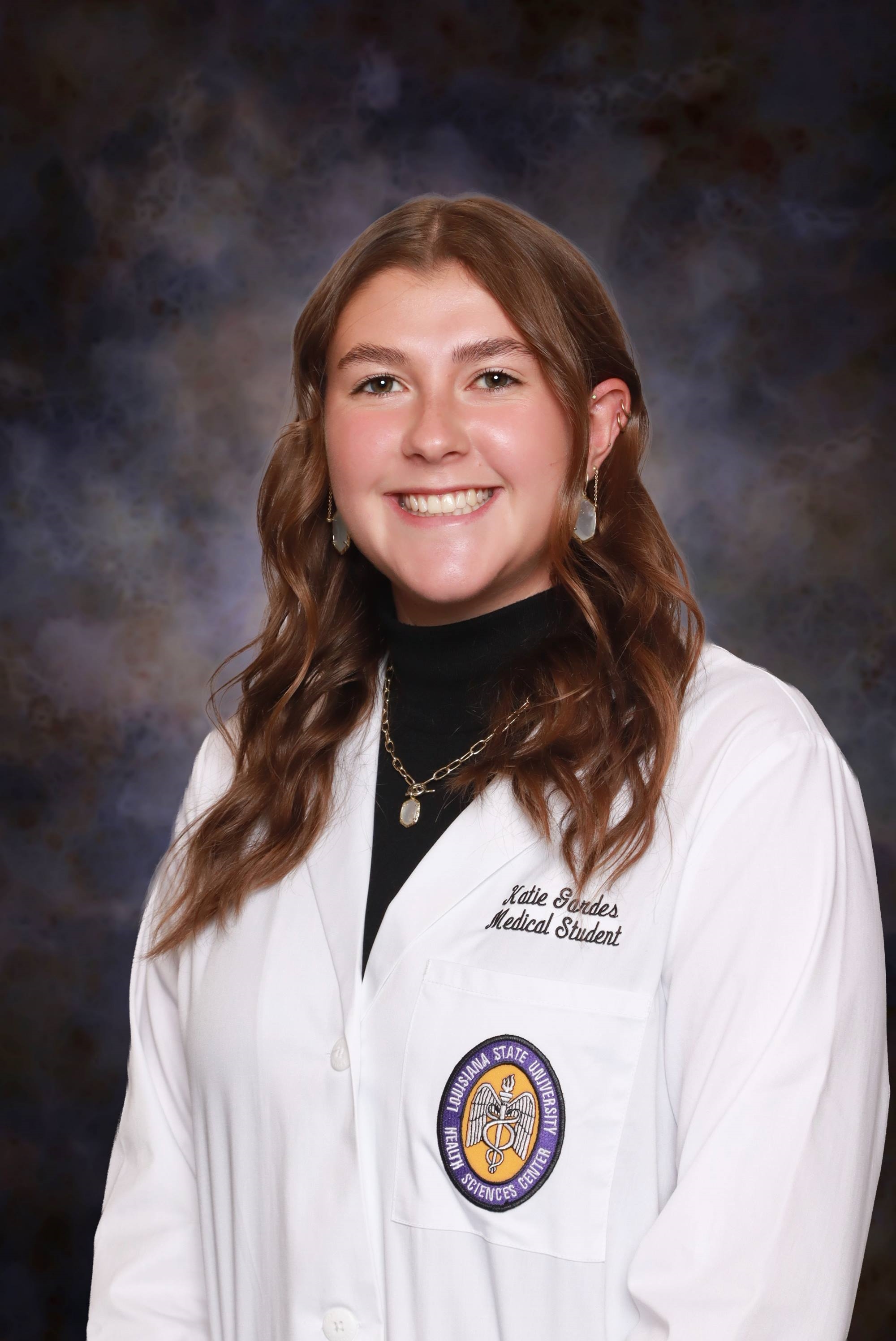 Professional medical school headshot showing a person wearing a white medical coat over a black high-neck top. Visible embroidered text on the coat includes the title “Medical Student,” and a Louisiana State University Health Sciences Center emblem is displayed on the chest pocket. The subject is wearing drop earrings, multiple ear piercings, and a necklace with a pendant. The portrait is set against a dark blue and gray mottled studio background with even, professional lighting.