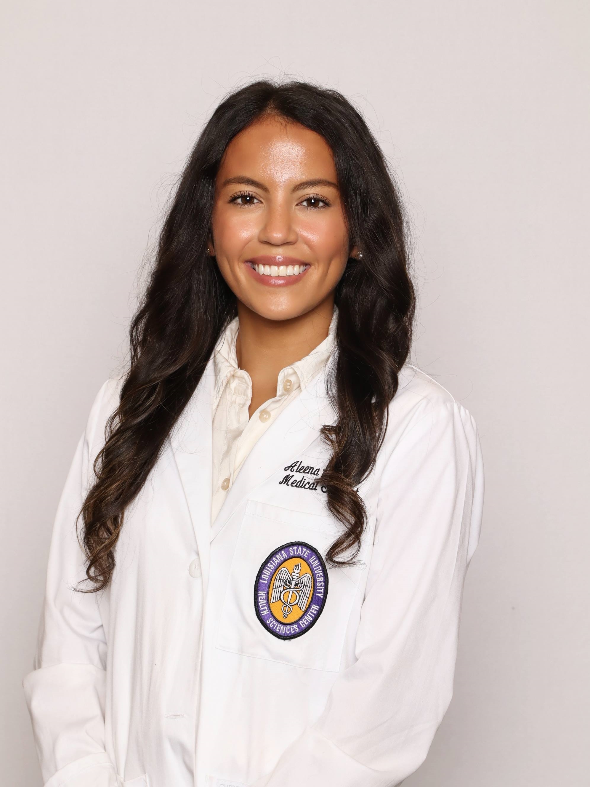 Professional medical school portrait showing a person wearing a white medical coat over a light-colored button-down blouse. Visible embroidered text on the coat includes the title “Medical Student,” and a Louisiana State University Health Sciences Center emblem is displayed on the chest pocket. The subject has long wavy hair and is facing the camera against a plain light gray background. The image is evenly lit and presented in a formal studio portrait style.