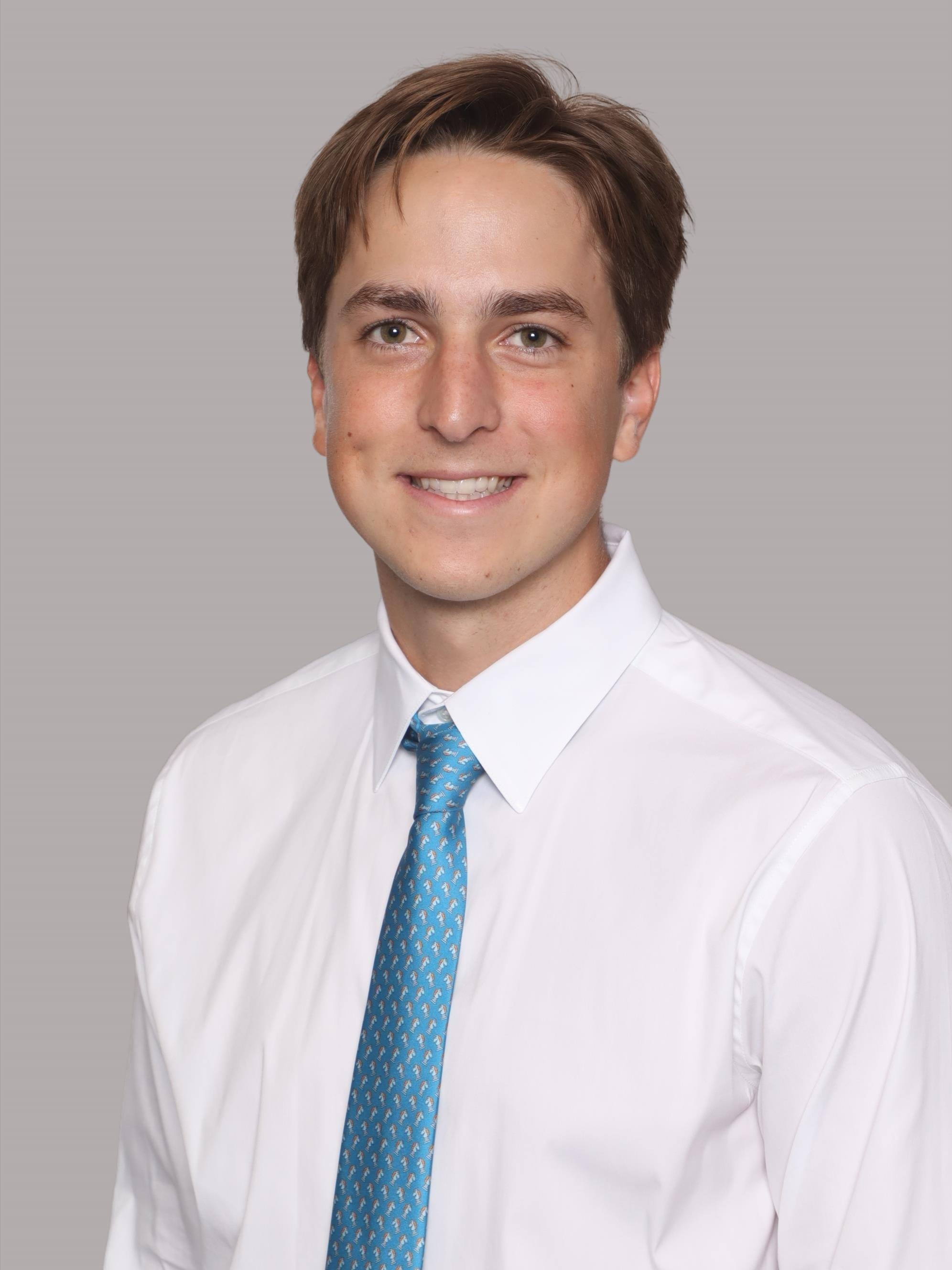 Professional headshot showing a person wearing a white dress shirt and a light blue patterned tie. The subject is facing the camera against a plain light gray background. The image is evenly lit and cropped from the chest up in a formal studio portrait style.