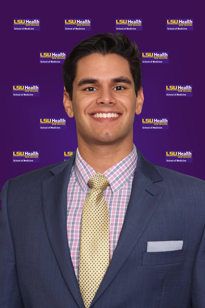 Professional headshot showing a person wearing a blue suit jacket, a pink-and-blue checkered dress shirt, a gold patterned tie, and a white pocket square. The subject is facing the camera against a purple LSU Health New Orleans School of Medicine branded background featuring multiple LSU Health New Orleans logos. The image is evenly lit and presented in a formal studio portrait style.