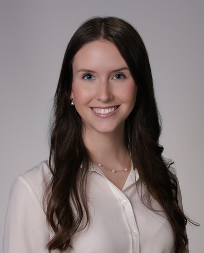 Professional headshot showing a person wearing a white button-down blouse and a delicate necklace. The subject is facing the camera against a plain light gray background. The image is evenly lit and cropped from the chest up in a formal studio portrait style.