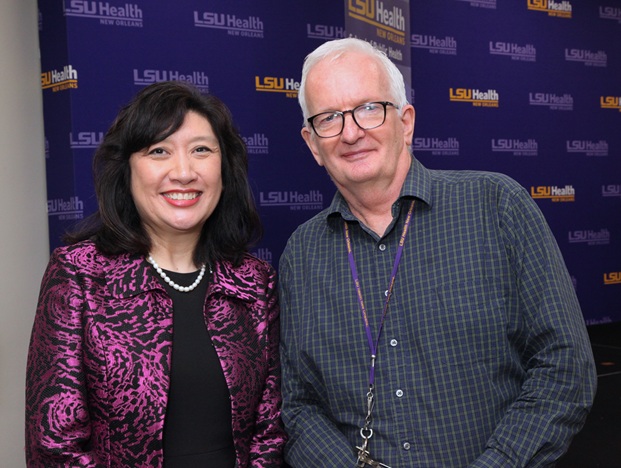 Two people pose side by side in front of a purple LSU Health New Orleans step-and-repeat backdrop displaying repeated LSU Health logos. One person wears a black outfit with a bright magenta patterned jacket and a pearl necklace. The other person wears glasses, a green-and-blue checkered button-down shirt, and a purple LSU Health lanyard with keys attached. The photo is taken indoors with both individuals facing the camera.