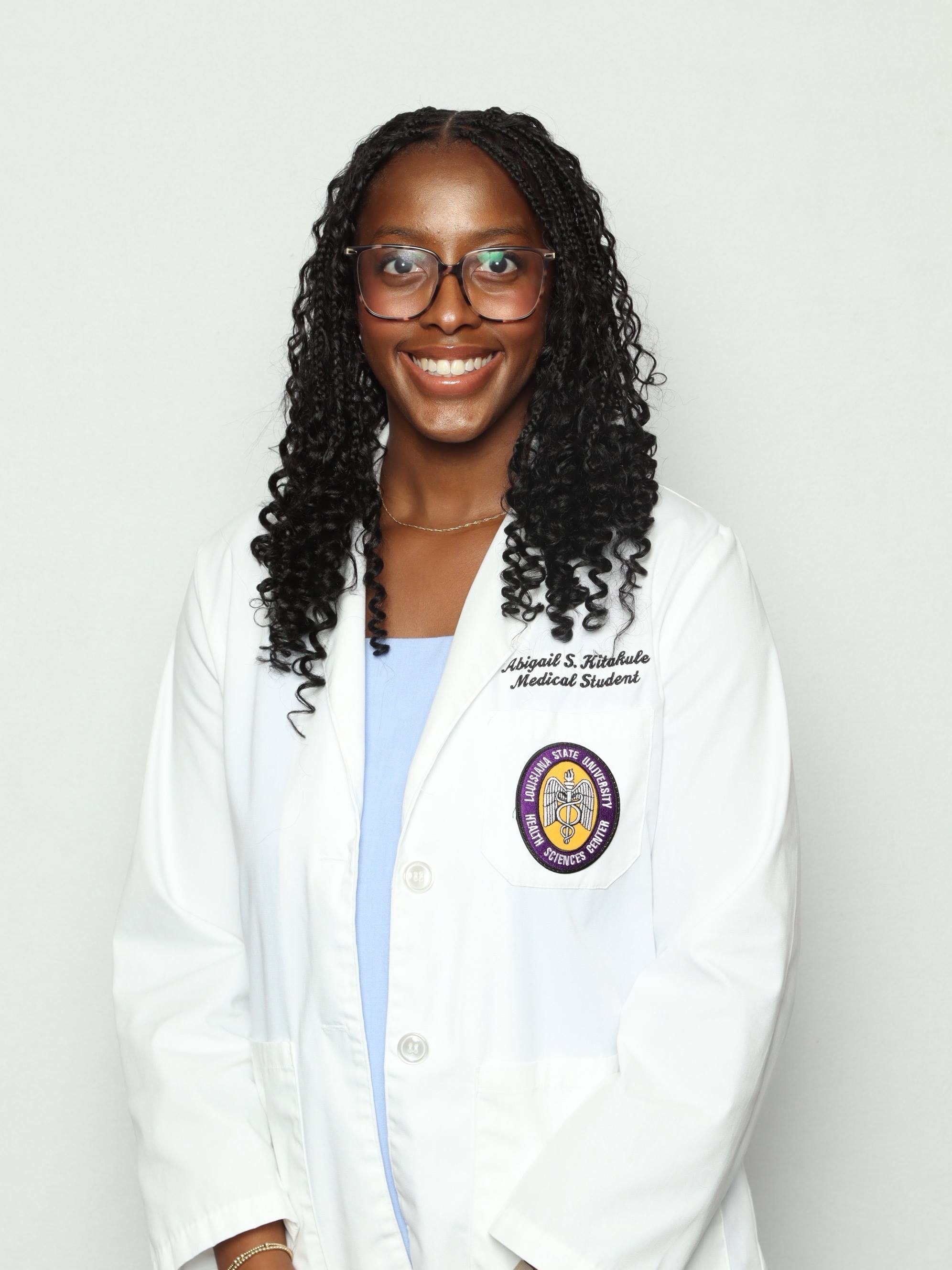 Professional medical school portrait showing a person wearing a white medical coat over a light blue top. Visible embroidered text on the coat includes the title “Medical Student,” and a Louisiana State University Health Sciences Center emblem is displayed on the chest pocket. The subject is wearing rectangular eyeglasses, a delicate necklace, and a bracelet. The portrait is set against a plain light gray background with even, professional lighting.