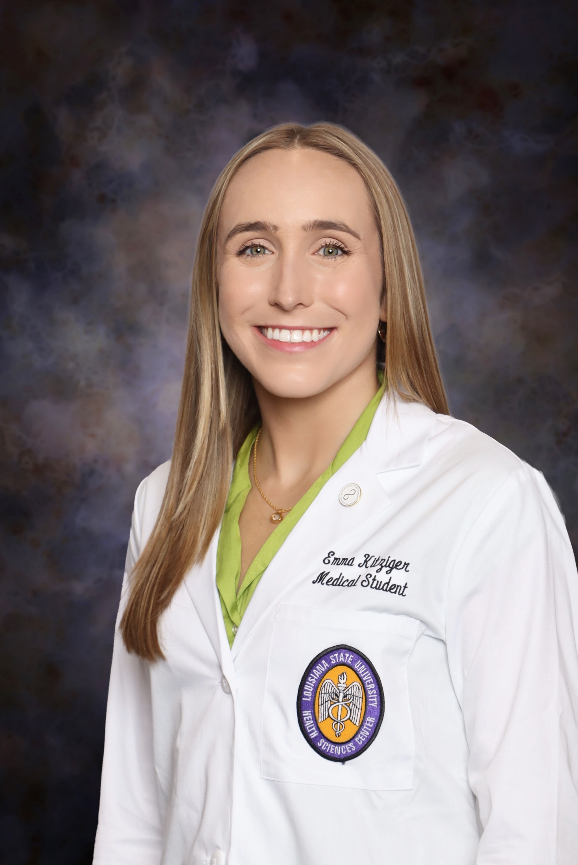Professional medical school headshot showing a person wearing a white medical coat over a light green collared shirt. Visible embroidered text on the coat includes the title “Medical Student,” and a Louisiana State University Health Sciences Center emblem is displayed on the chest pocket. The subject is facing the camera against a dark blue and gray mottled studio background with even, professional lighting.