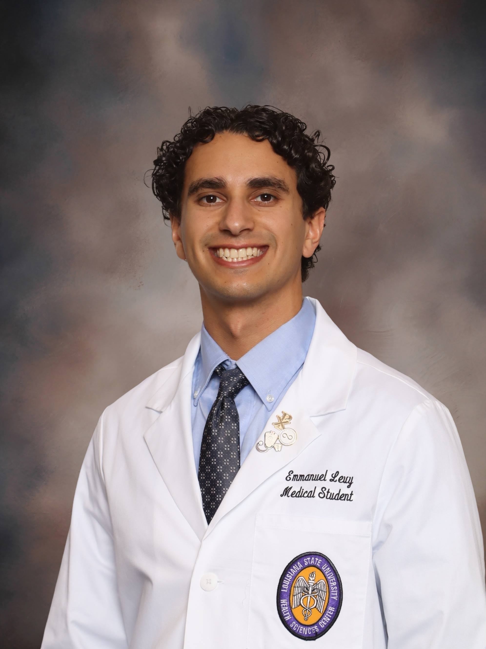 Professional medical school headshot showing a person wearing a white medical coat over a light blue dress shirt and patterned tie. Visible embroidered text on the coat includes the title “Medical Student,” and a Louisiana State University Health Sciences Center emblem is displayed on the chest pocket. A decorative lapel pin is attached to the coat. The subject is facing the camera against a softly blended gray and brown studio background with even, professional lighting.