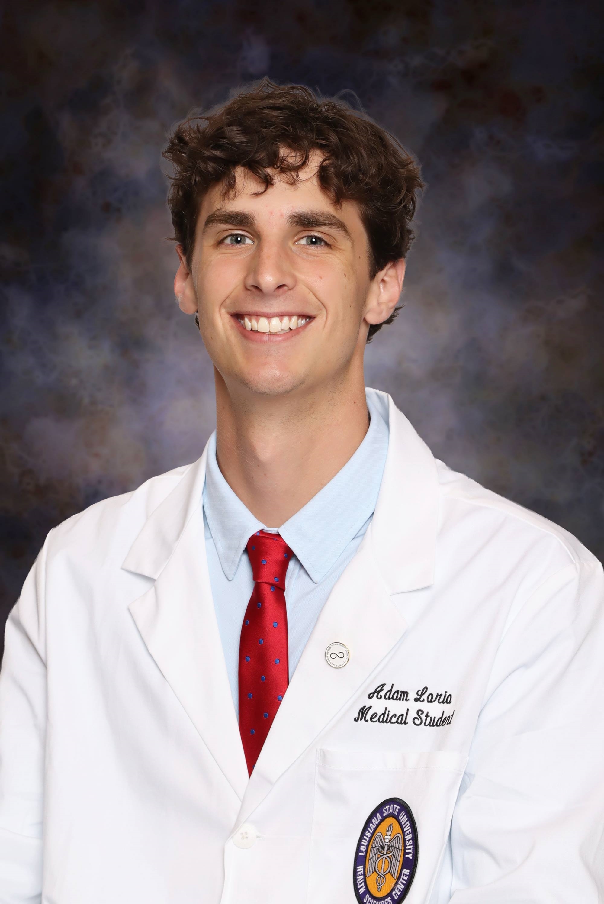 Professional medical school headshot showing a person wearing a white medical coat over a light blue dress shirt and a red tie with a small blue pattern. Visible embroidered text on the coat includes the title “Medical Student,” and a Louisiana State University Health Sciences Center emblem is displayed on the chest pocket. A circular lapel pin is attached to the coat. The portrait is set against a dark blue and gray mottled studio background with even, professional lighting.