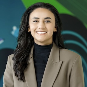 Professional headshot of a person wearing a tan blazer over a black turtleneck, standing in front of a colorful abstract background.