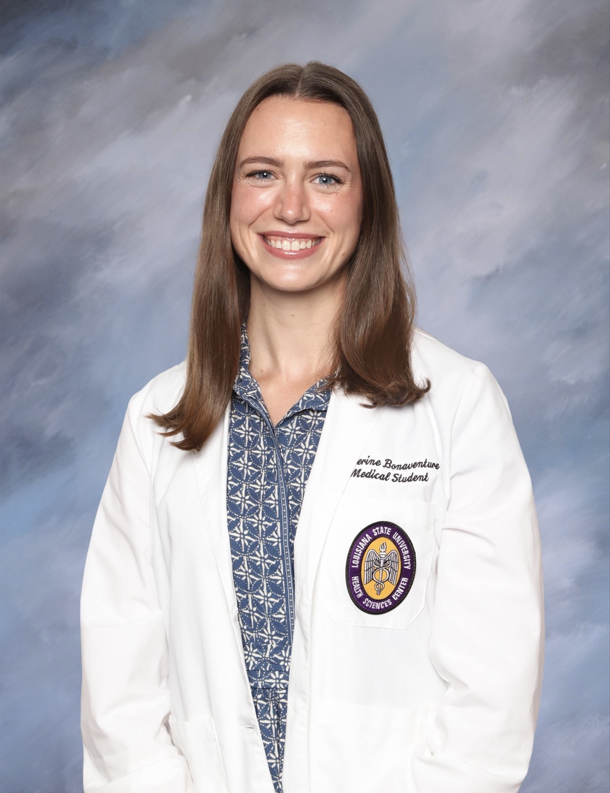 Professional medical school portrait of a person wearing a white medical coat over a blue patterned shirt against a light blue-gray studio background.