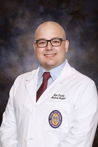 Professional medical school portrait of a person wearing a white medical coat, light blue shirt, and dark red tie against a studio background.