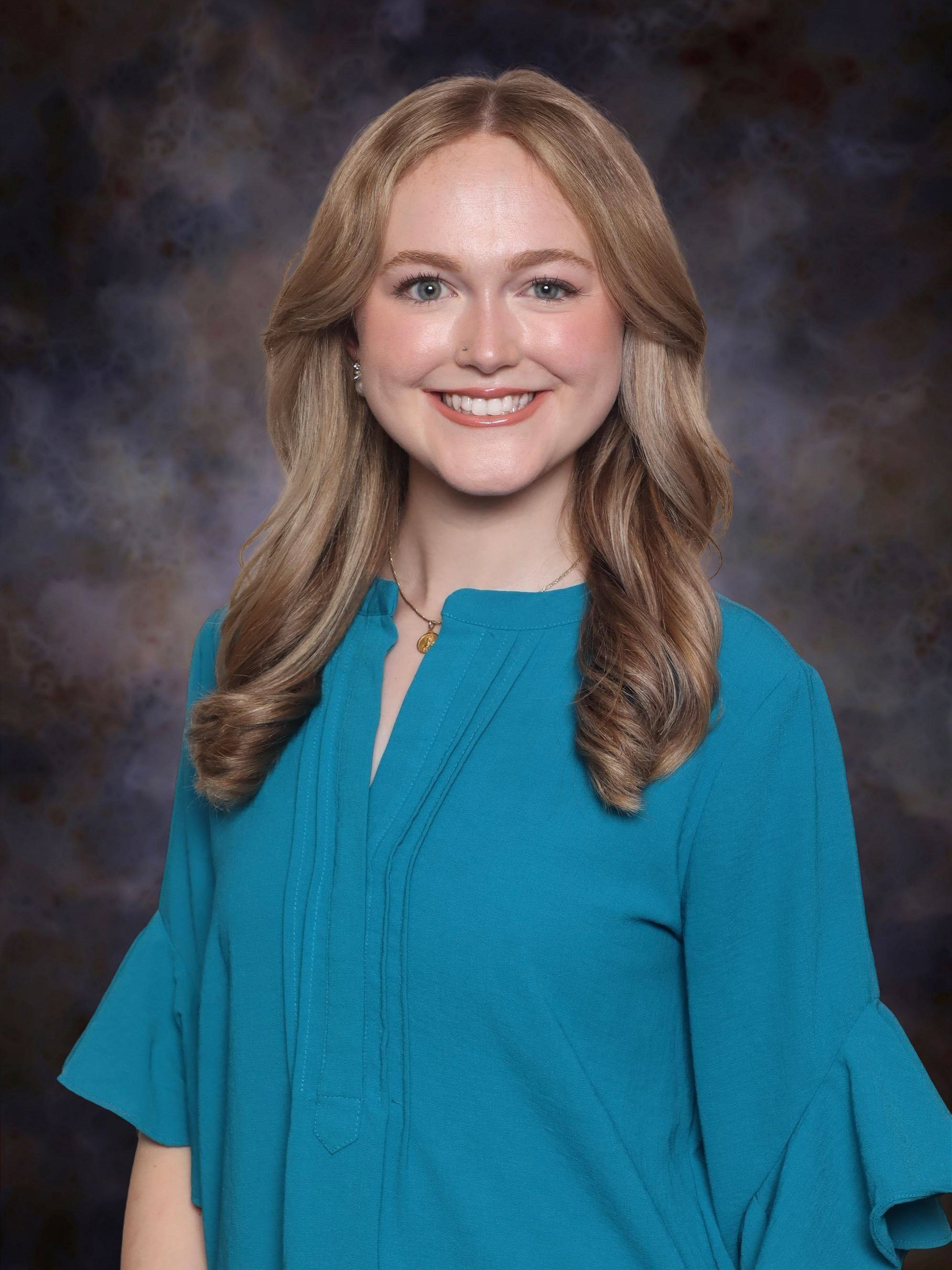 Professional studio portrait of a person wearing a teal blouse against a mottled dark blue and gray background.