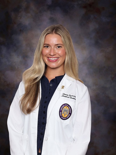 Professional medical school portrait of a person wearing a white medical coat over a navy blue blouse against a dark blue-gray studio background.