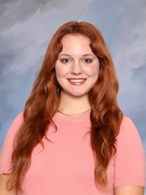 Professional studio portrait of a person with long wavy red hair wearing a light pink short-sleeved top against a light blue-gray background.