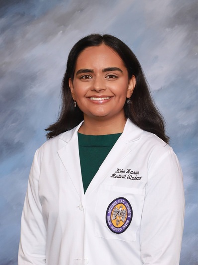 Professional medical school portrait of a person wearing a white medical coat over a green top against a light blue and gray studio background.