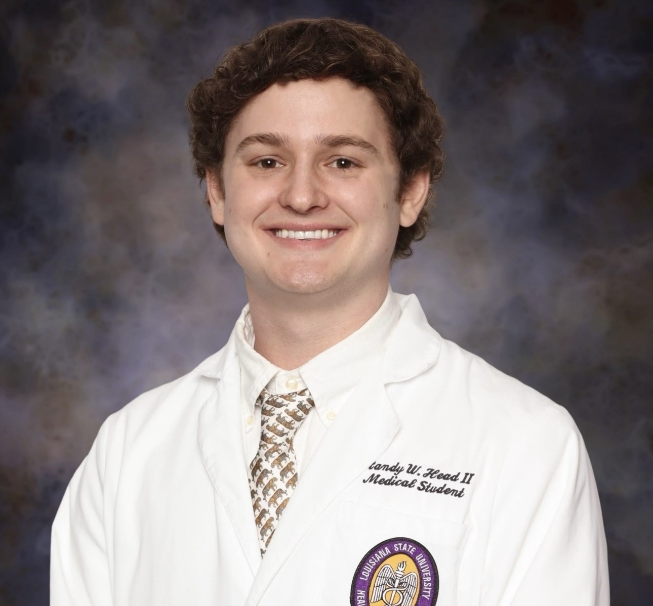 Professional portrait of a person wearing a white medical coat and patterned tie against a studio backdrop.