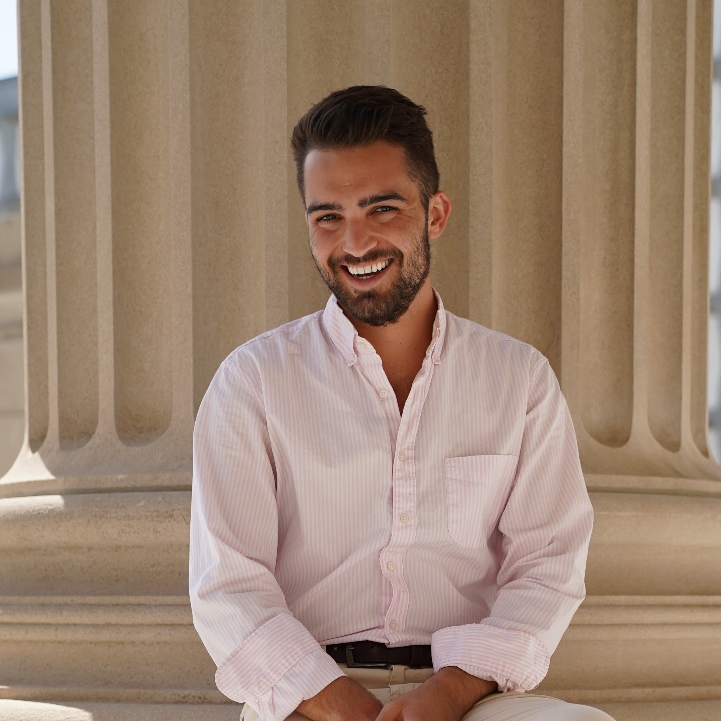Professional outdoor portrait of a person wearing a light pink button-down shirt, seated in front of large stone columns.