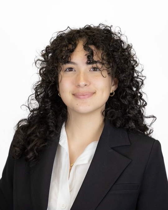 Professional headshot of a person with curly dark hair wearing a black blazer over a white collared shirt against a light gray background.